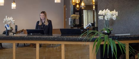 Una mesa de recepción con una recepcionista, una barra de madera y un jarrón con flores blancas.