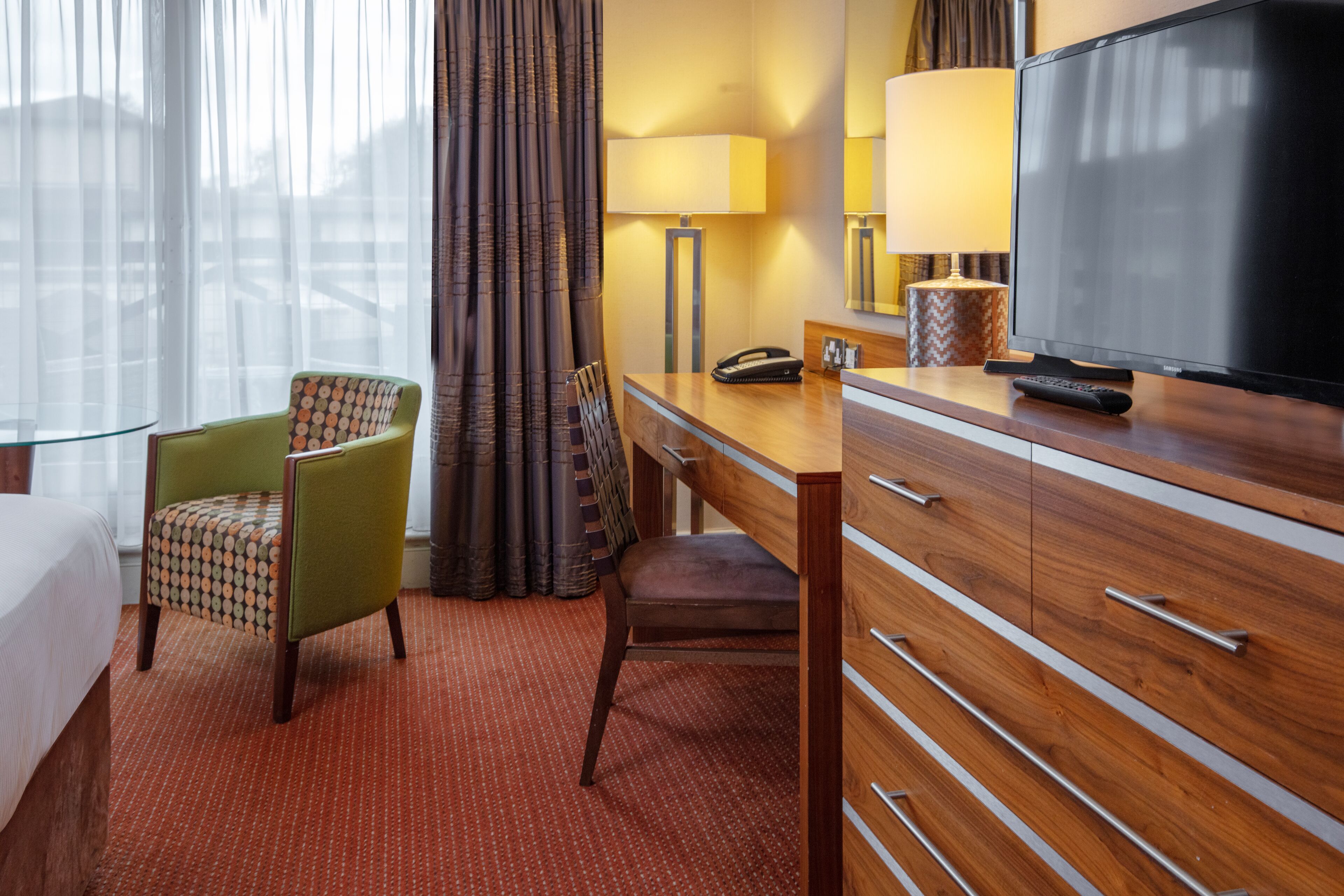 A hotel room with a bed, a desk with a lamp, a television, a chair, and a dresser.