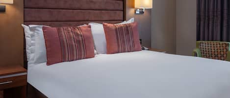 Una habitación de hotel con una cama grande, cabecera, mesita de noche, lámpara y una silla.