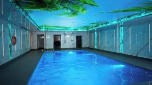 Una amplia piscina interior con superficie de agua azul cristalina, rodeada de paredes con franjas de luz verticales y un techo con diseño de palmeras.