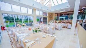 Un amplio comedor interior con varias mesas dispuestas para un evento formal, ventanas grandes y un techo alto.