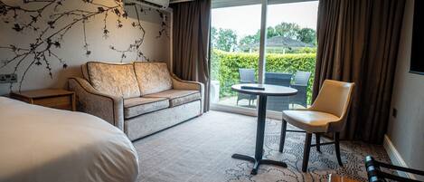 A hotel room with a bed, a sofa, a chair, a small table, and a view of the outdoors through a sliding door.