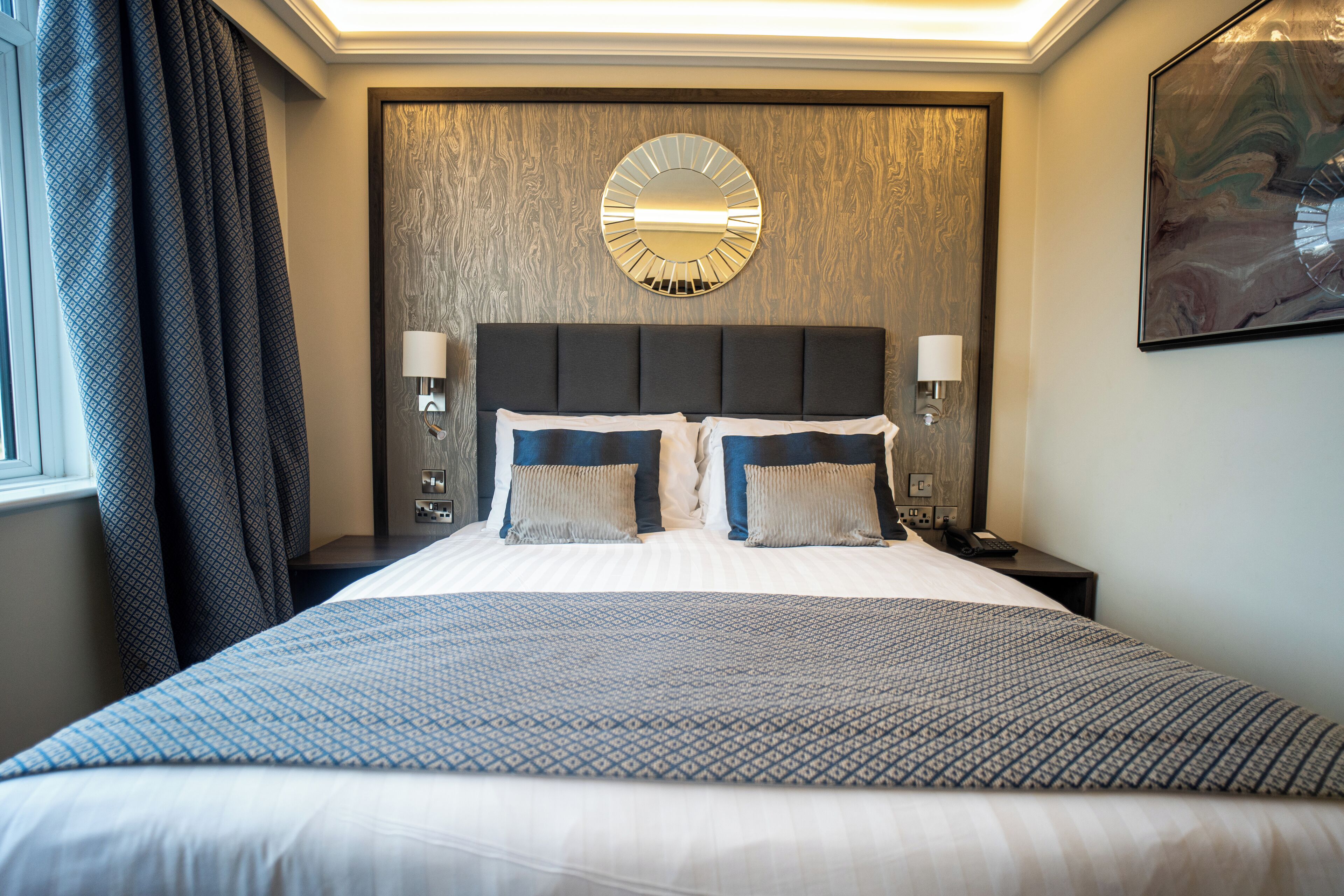 A hotel room with a large bed, two bedside lamps, a round mirror on the wall, and a painting above the bed.