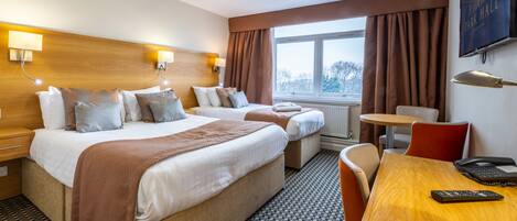 A hotel room with two beds, a wooden headboard, a small table, a chair, a TV, and a window with curtains.