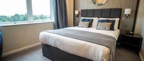 A hotel room with a large bed, a round wall clock, a nightstand with a lamp, and a view of trees through the window.