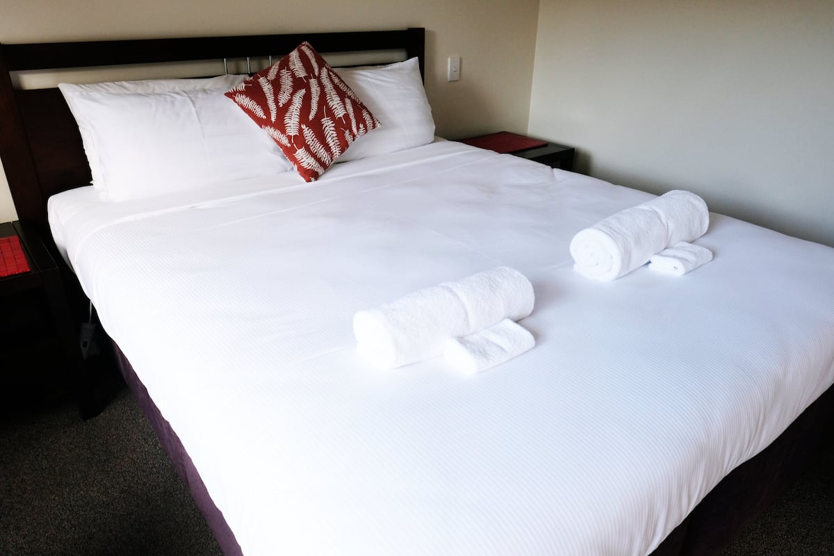 A neatly made bed with white linens and a red patterned pillow, accompanied by two rolled towels.