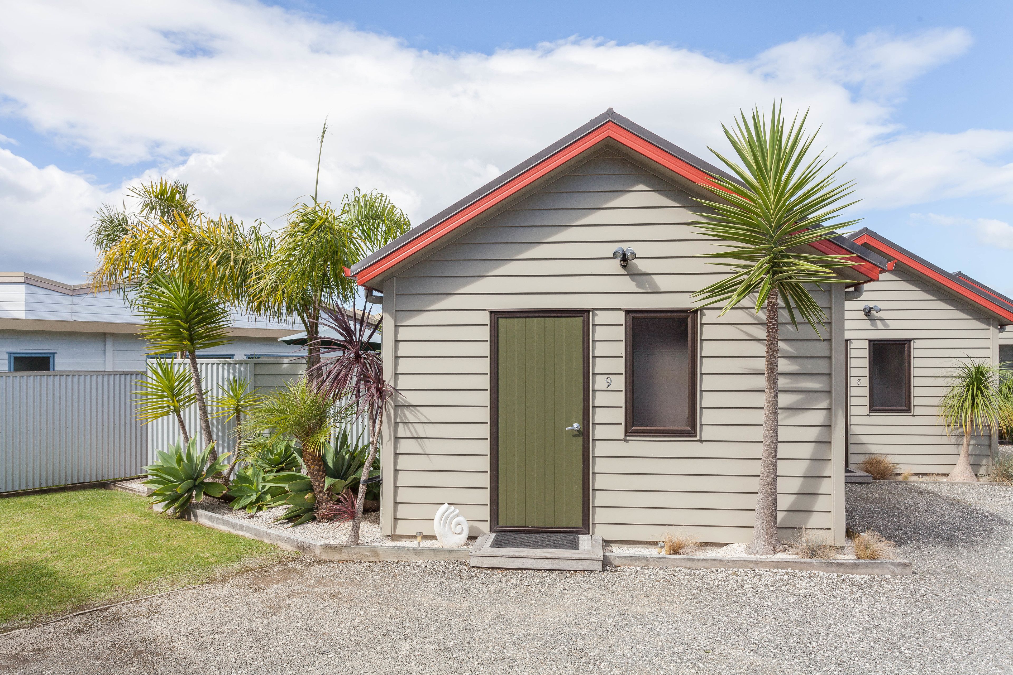 A single-story house with a green door, a red roof, and a small front yard with palm trees and a white fence.