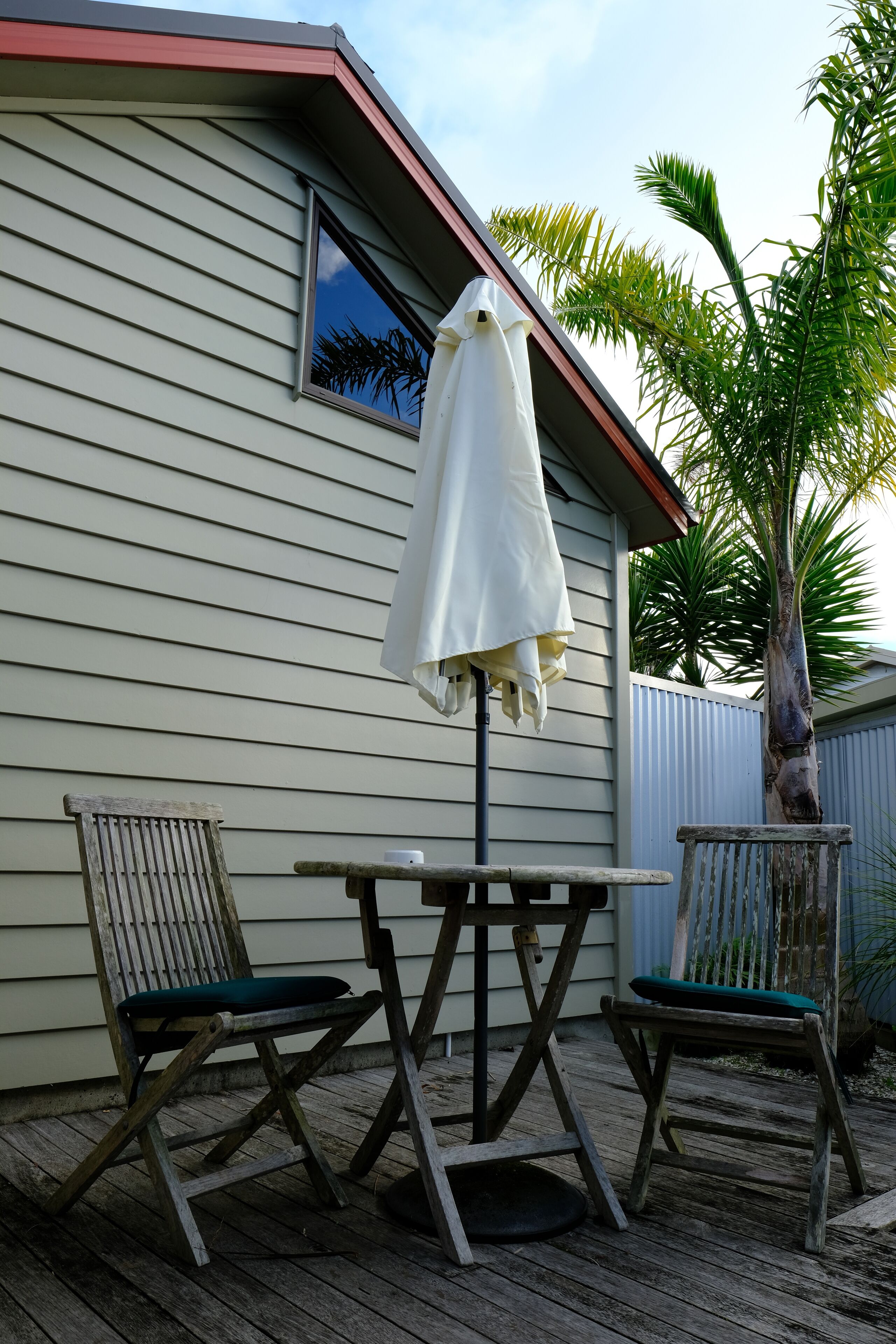 Un patio in legno con due sedie e un tavolo, un ombrellone bianco e una casa con rivestimento e una finestra.