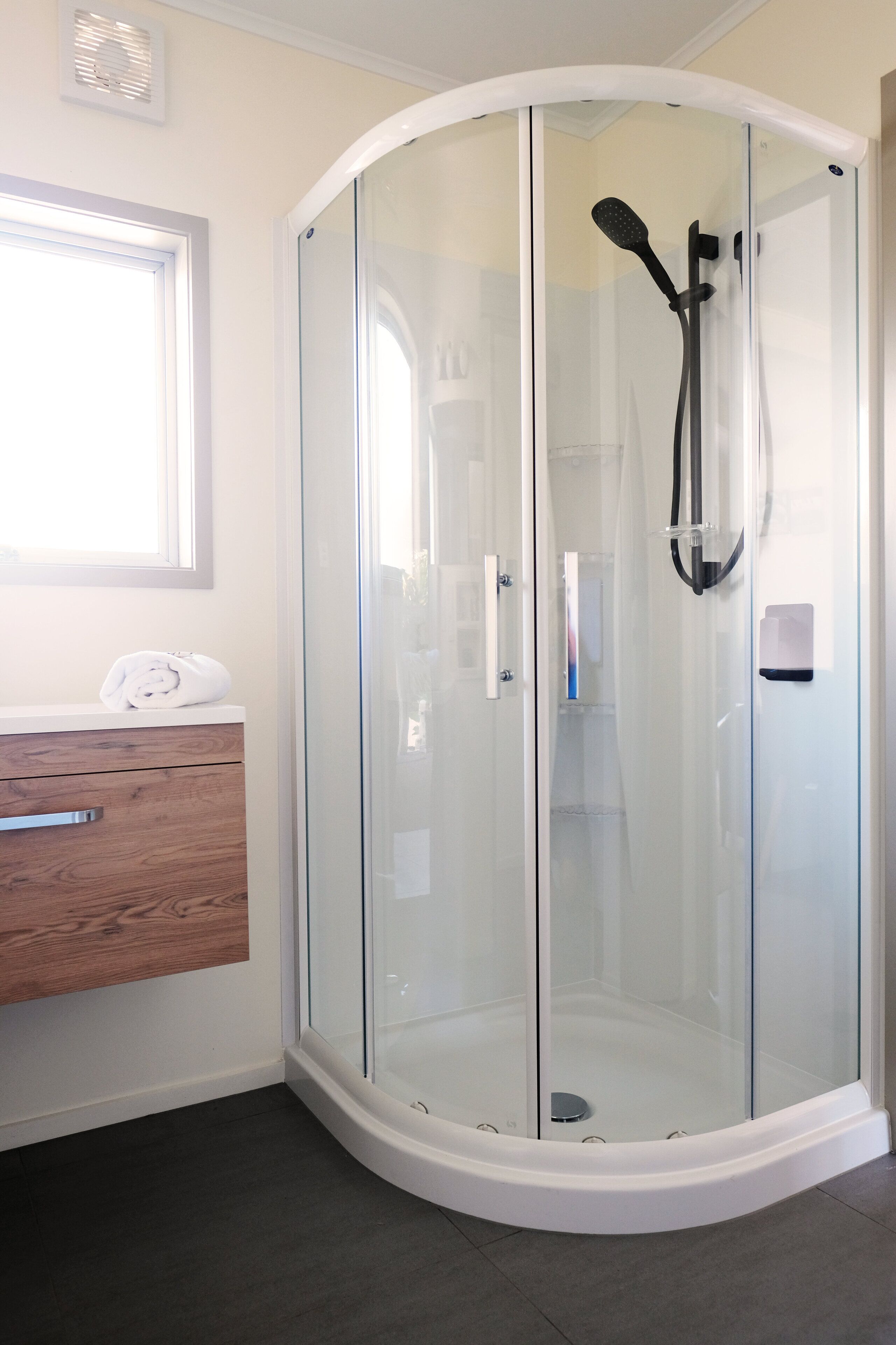A glass-enclosed shower with a handheld showerhead, a wooden vanity with a towel, and a window above the vanity.