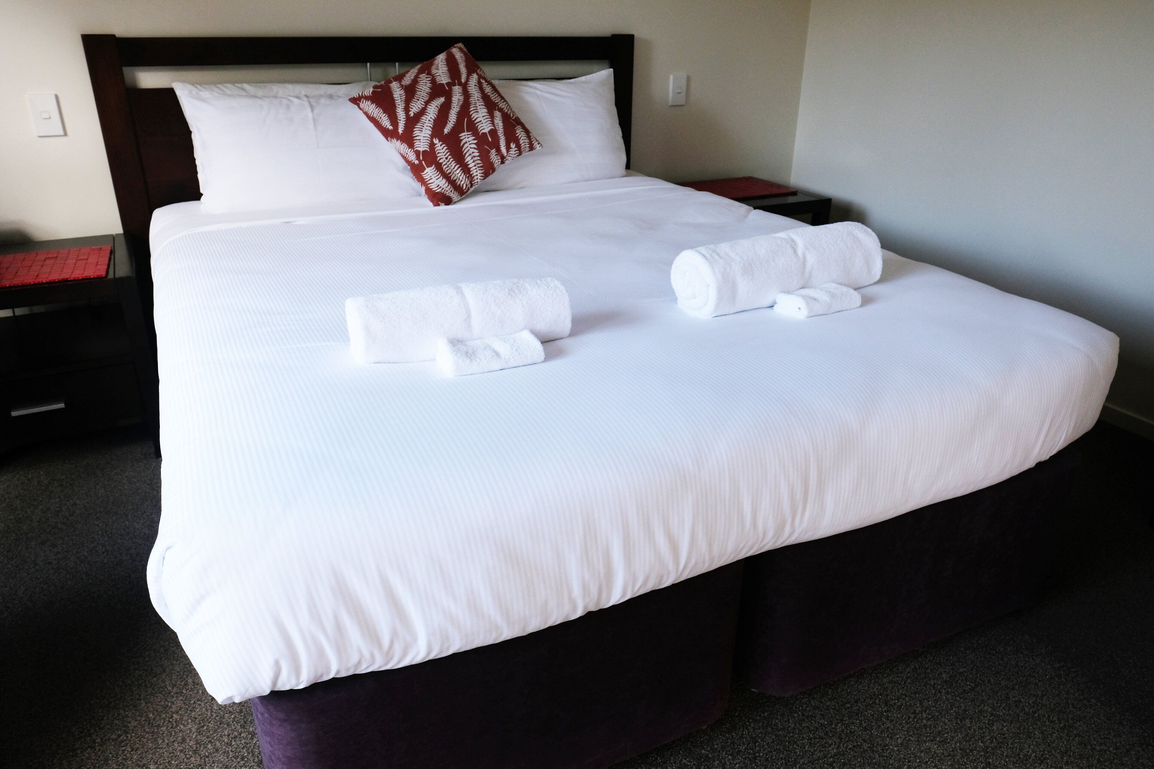 A neatly made bed with white linens, two rolled towels, and a decorative pillow.