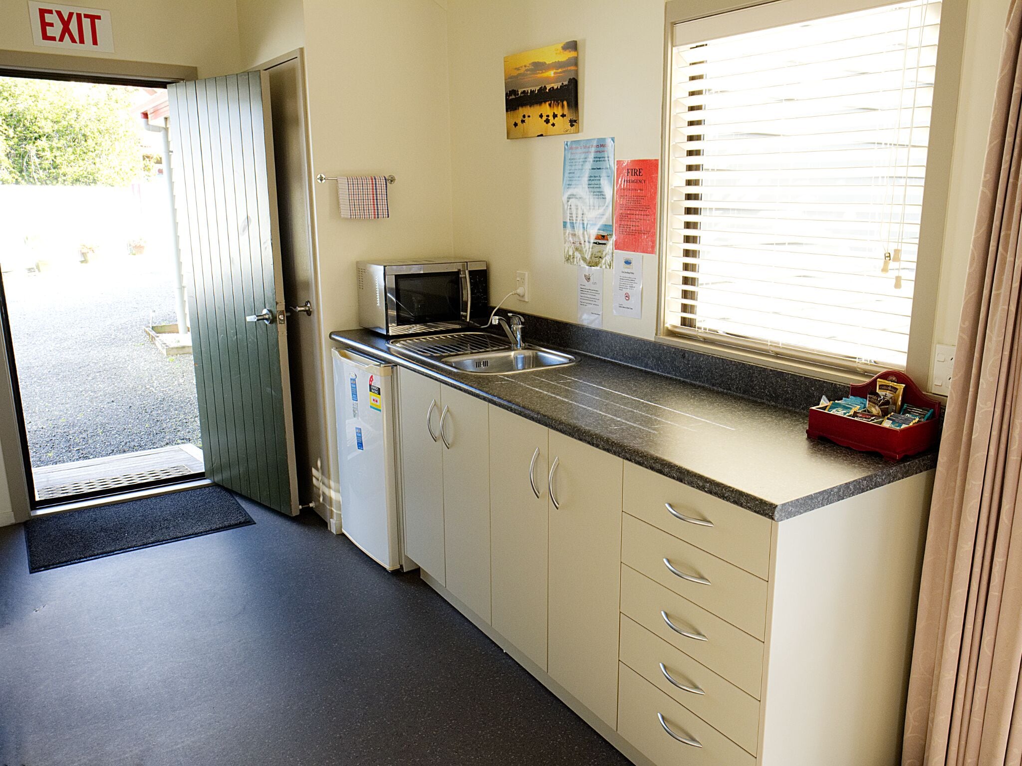 Una pequeña kitchenette con microondas, lavabo y armarios. Hay una ventana con persianas y una puerta señalizada con 