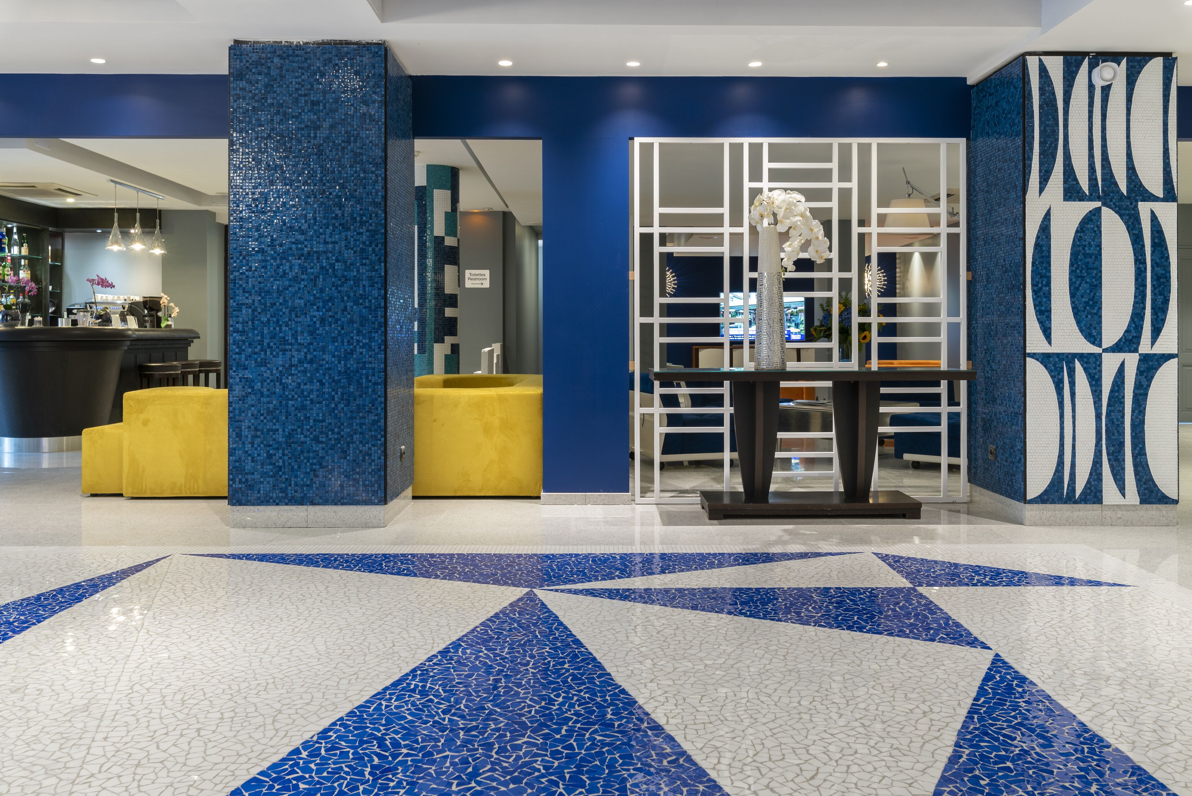 Un vestíbulo con suelo de mosaico azul y blanco, una zona de estar amarilla y un mostrador de recepción con barra.