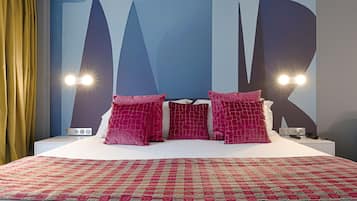 A bed with a red and beige patterned bedspread, a white headboard, and three pink pillows. There are two bedside lamps with lights on, and a nightstand with a phone and a remote control.