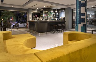 Een moderne bar met een gebogen toonbank, diverse krukken en een zithoek met gele banken.