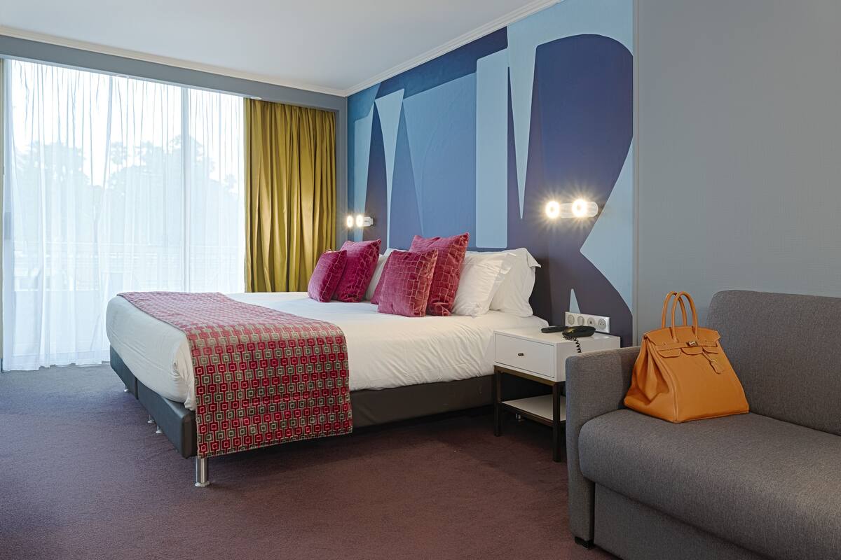 A hotel room with a bed, a grey sofa, a nightstand, and a wall with abstract art.