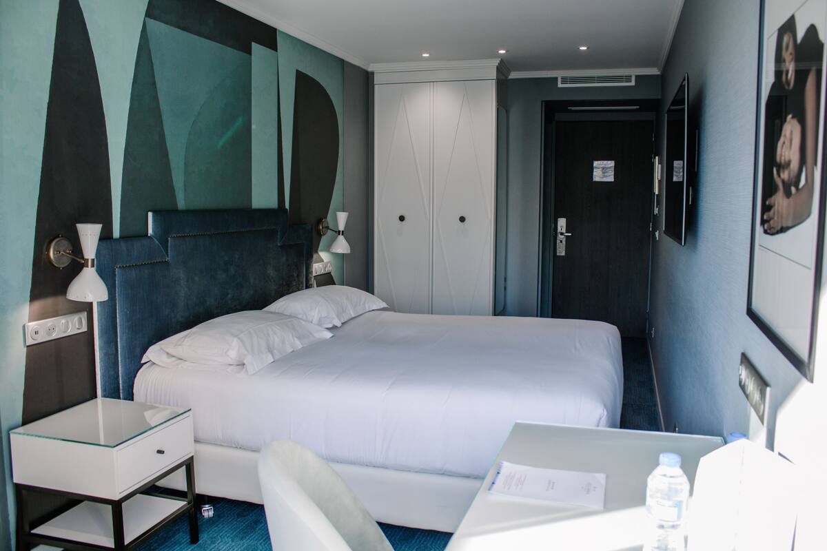 A hotel room with a bed, bedside table, lamp, and a desk with a chair.
