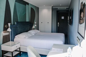 Hotellihuone, jossa on sänky, yöpöytä, lamppu ja työpöytä tuolilla.