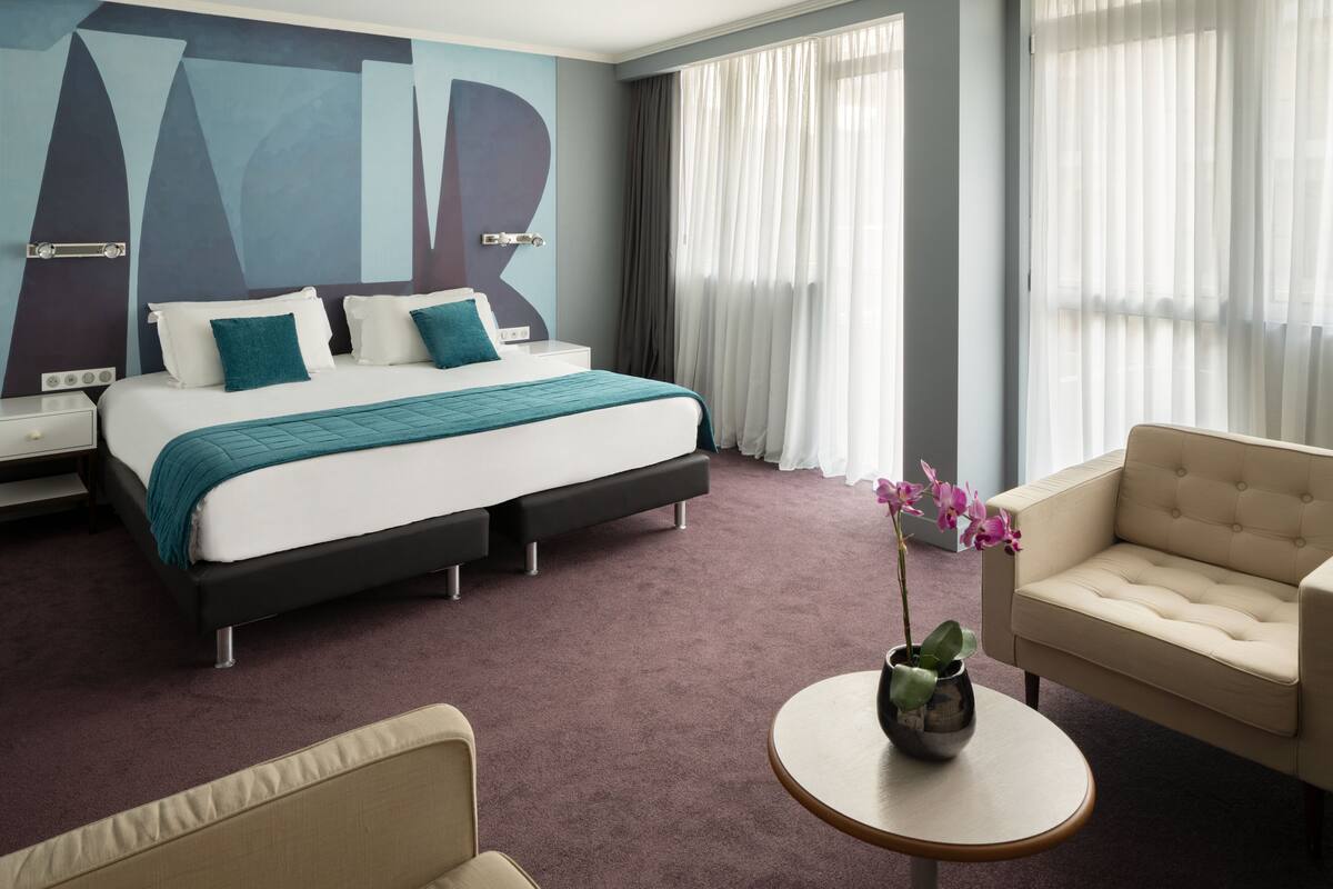 A hotel room with a bed, a chair, a small table, and a vase with flowers.