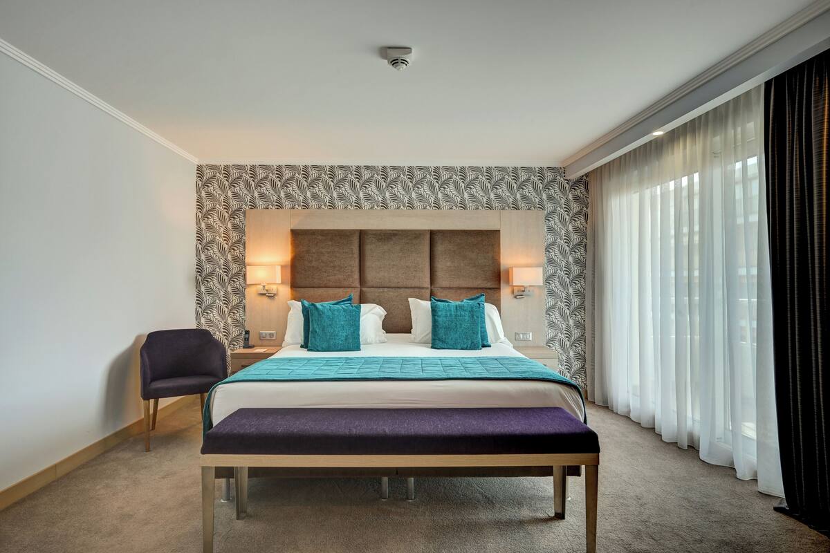 A hotel room with a large bed, a bench, a chair, and a view of the city through sheer curtains.