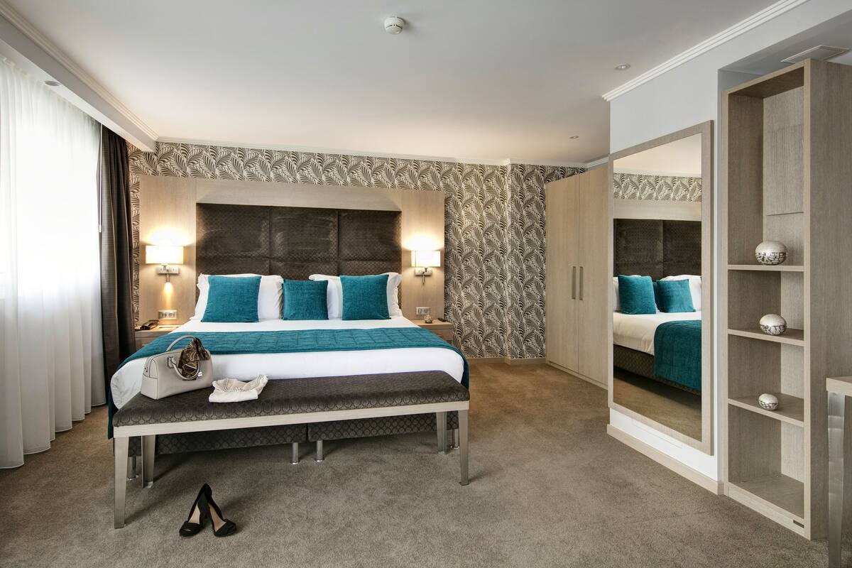A hotel room with a large bed, bedside lamps, a bench, a mirror, and a wardrobe.