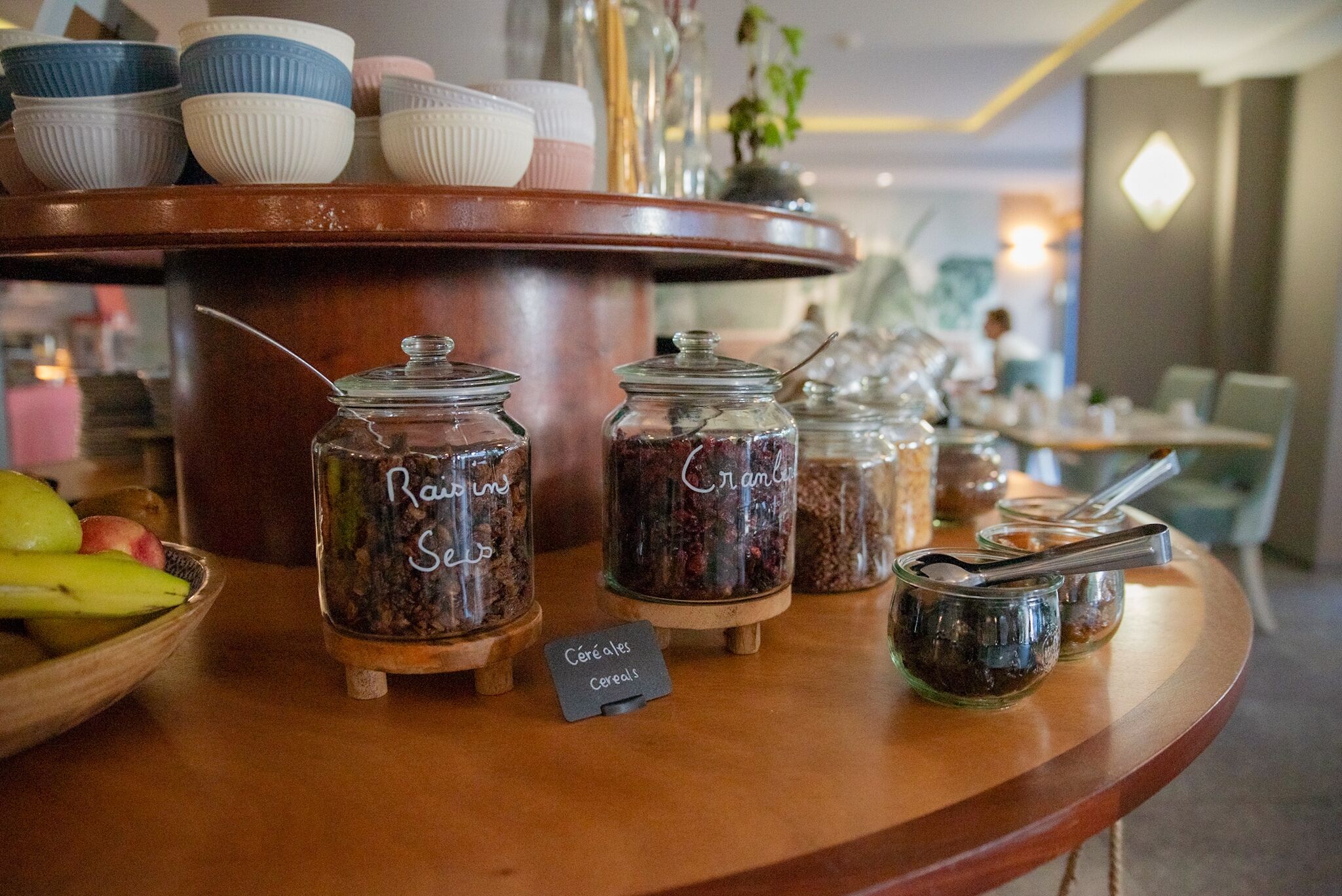 A breakfast buffet with various cereal jars labeled 'Raisin' and 'Cranberry' on a wooden counter.
