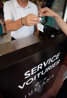Una persona entregando un dispositivo a otra persona en un mostrador con la leyenda 'SERVICE VOITURIER'.
