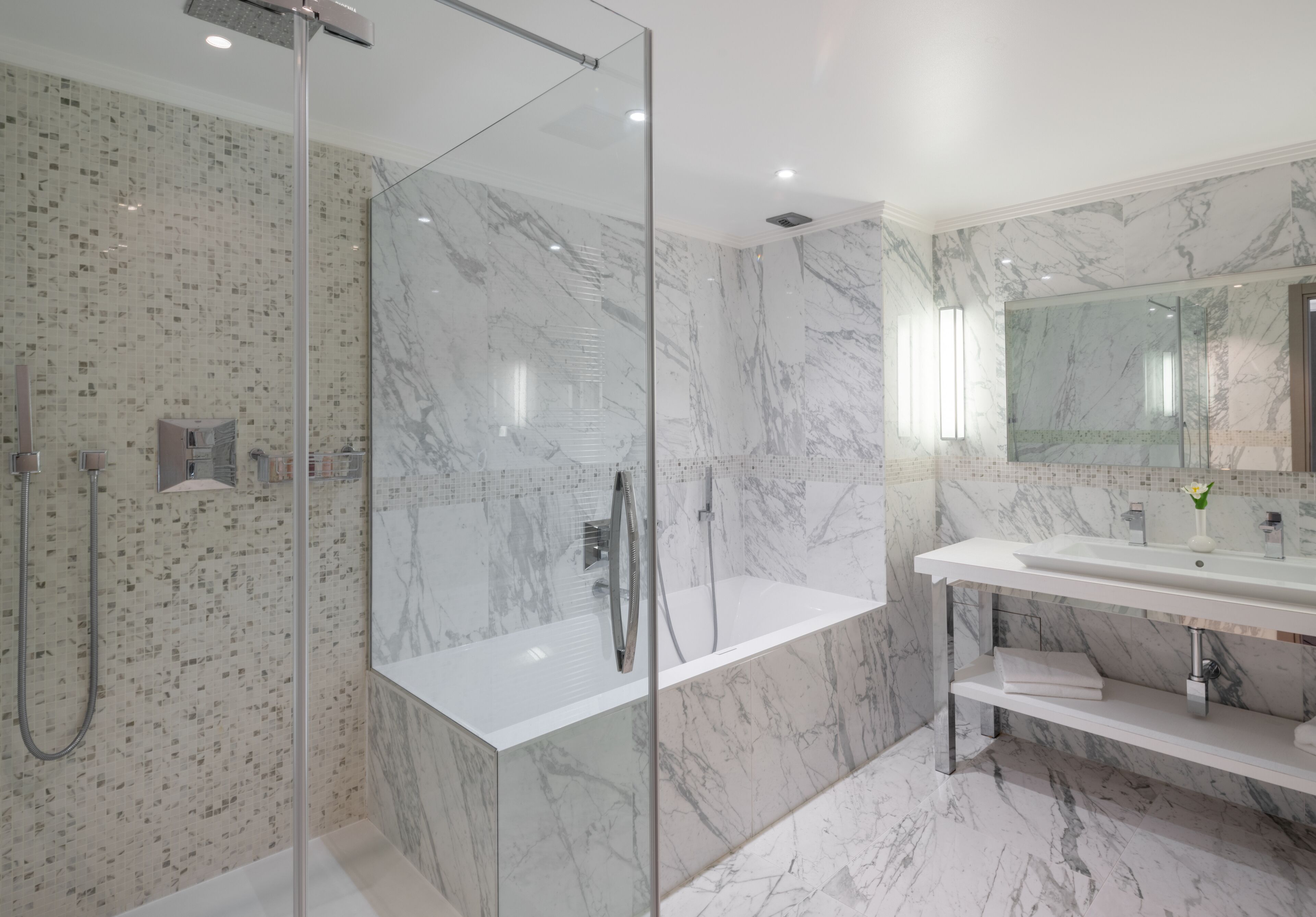 A modern bathroom with a glass-enclosed shower, a marble-tiled wall, a white sink, and a large mirror.