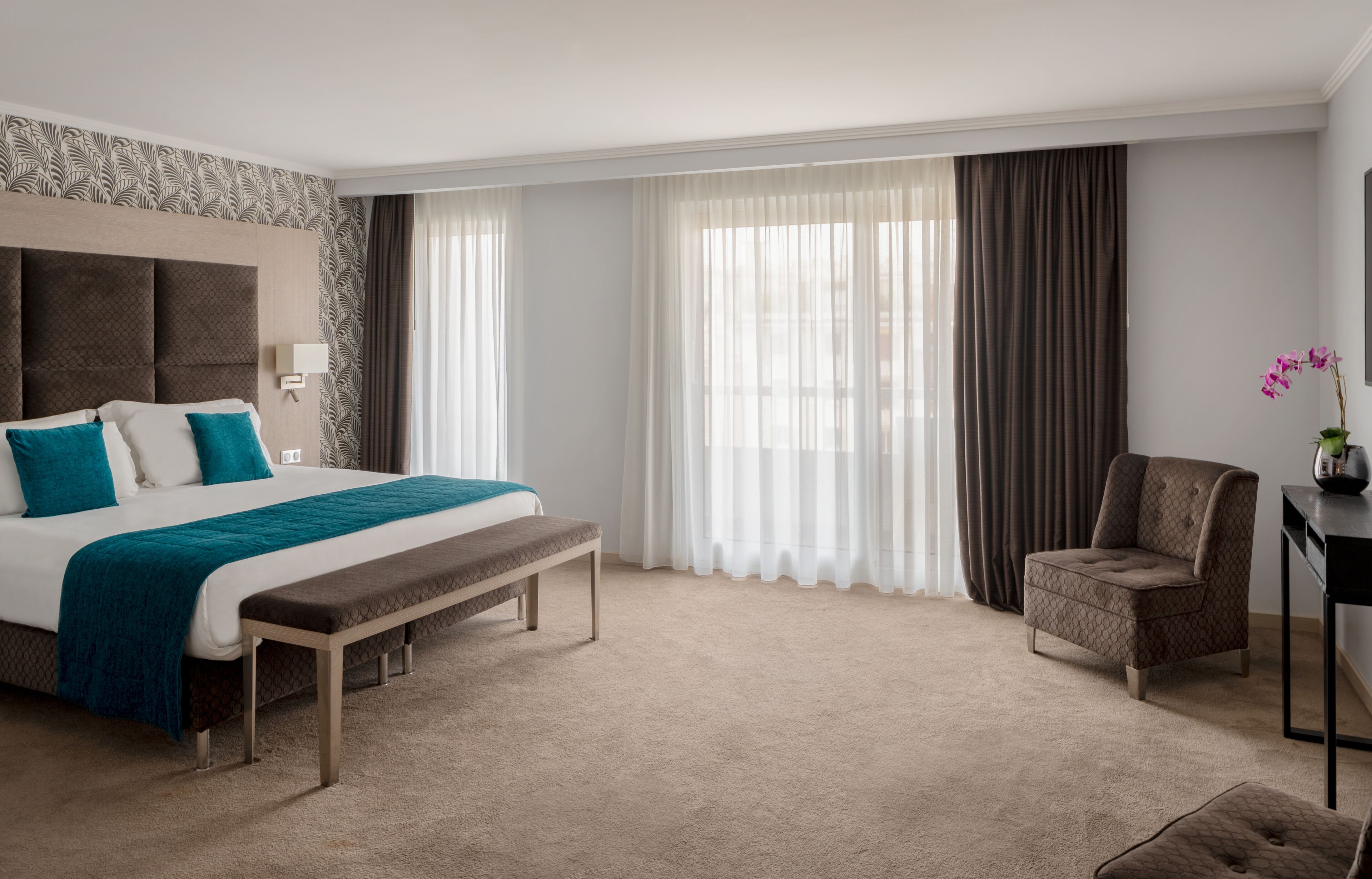 A hotel room with a large bed, a bench, a chair, a desk, and a window with curtains.