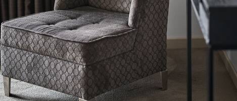 Un sillón moderno con un estampado geométrico, ubicado en una habitación con una cortina oscura y una planta en una mesita auxiliar.