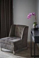 A modern armchair with a geometric pattern, positioned in a room with a dark curtain and a potted plant on a side table.
