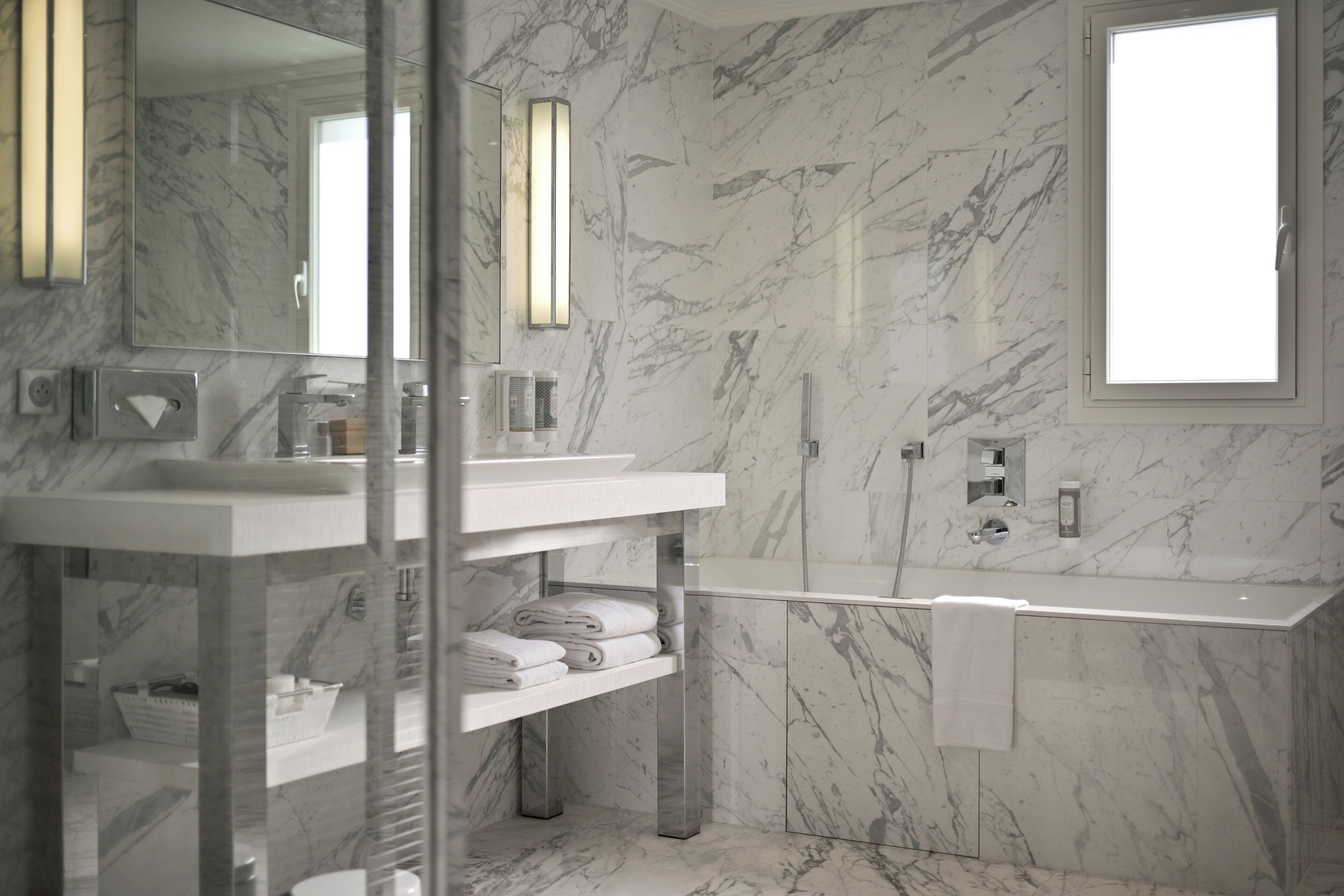 A modern bathroom with a marble floor, a large mirror, a white sink, and a glass-enclosed shower area.