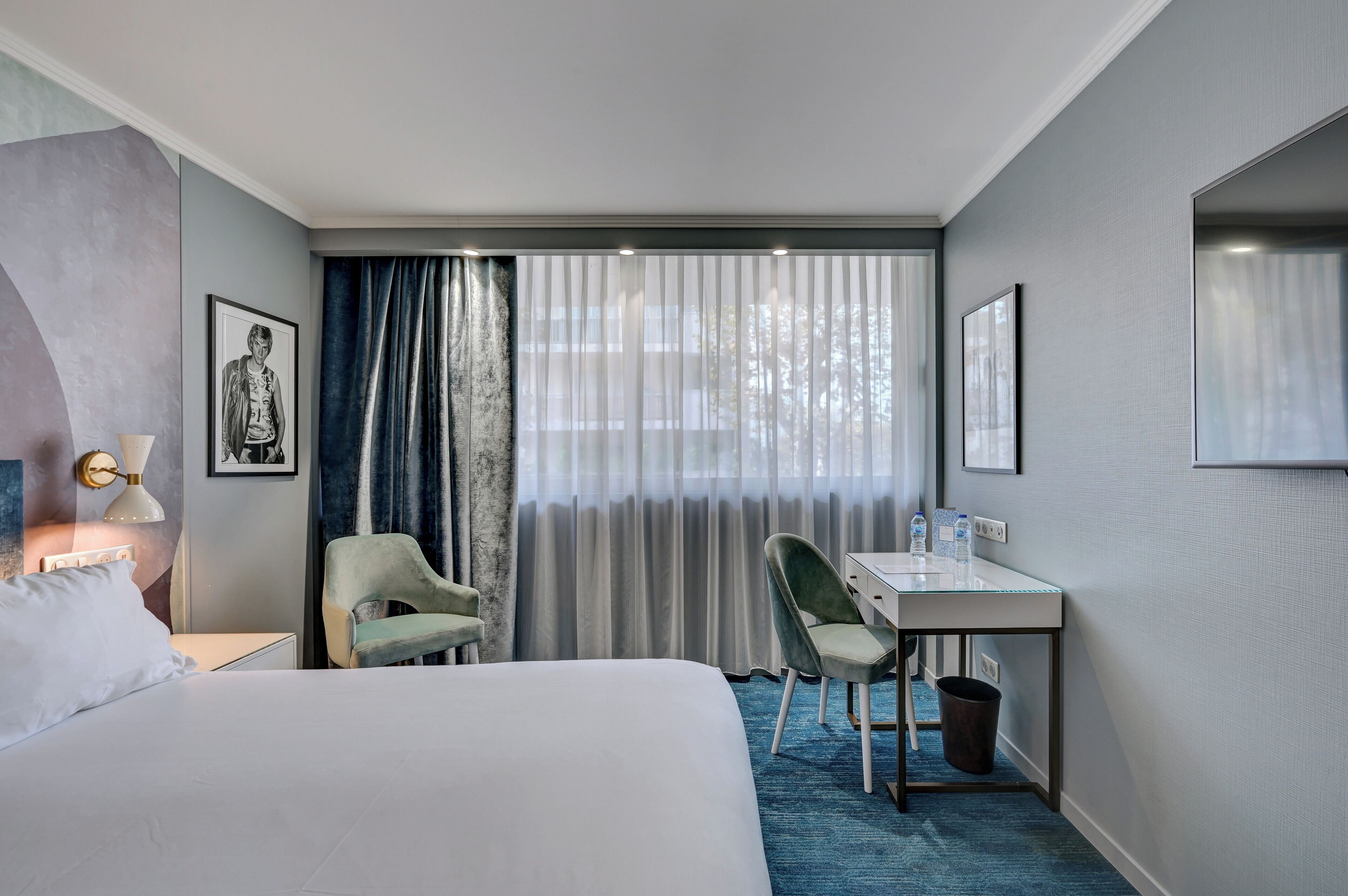 Habitación de hotel con cama, escritorio, silla y un ventanal con cortinas.