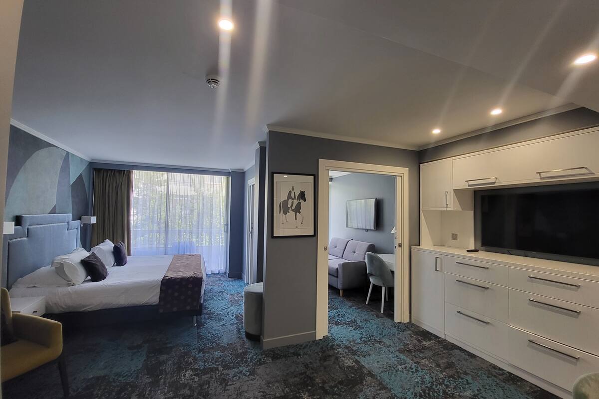 A modern hotel room with a bed, a sofa, a TV, and a kitchenette.
