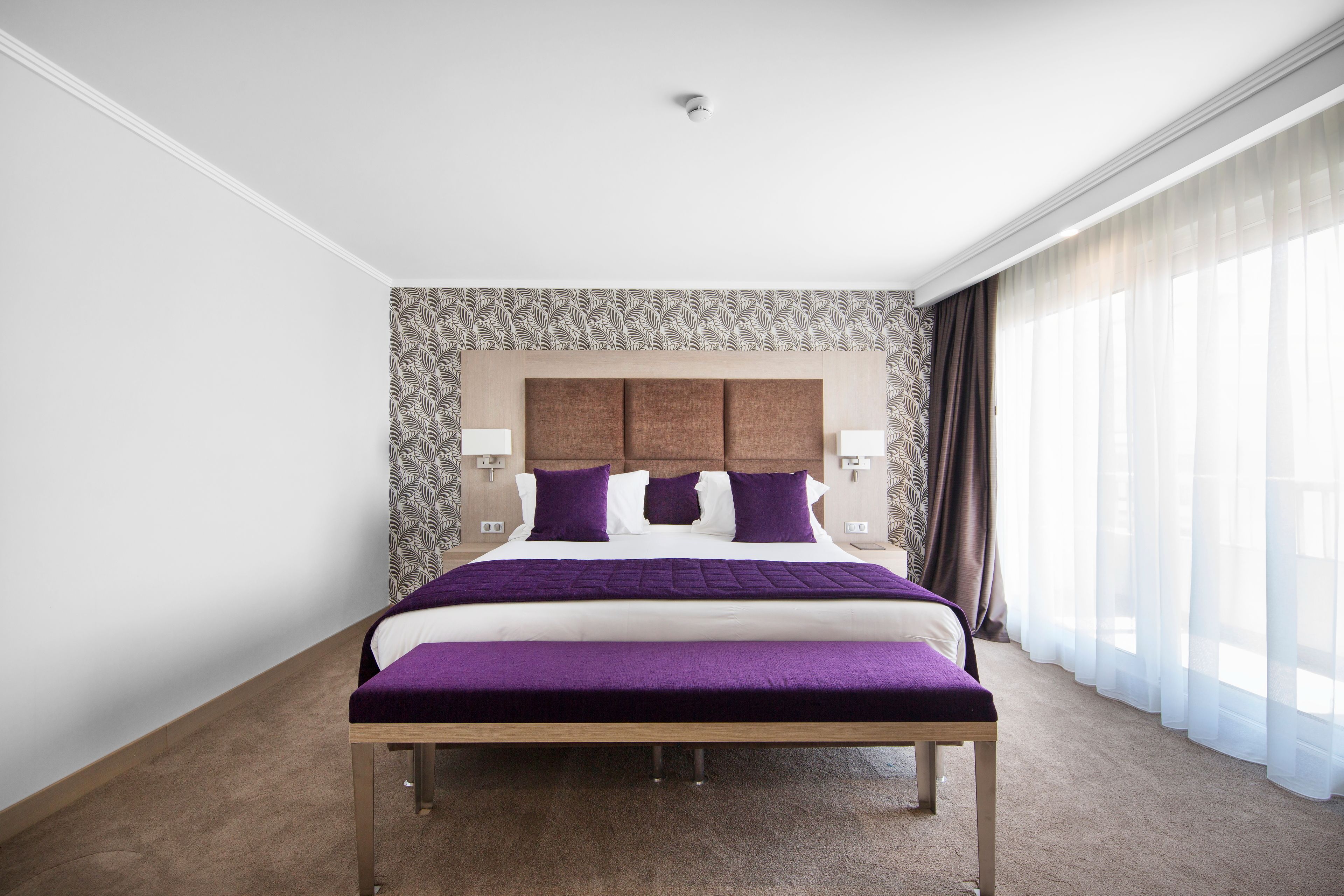 Una habitación de hotel con una cama grande, un cabecero con papel tapiz estampado, una mesita de noche con una lámpara y cortinas translúcidas en la ventana.