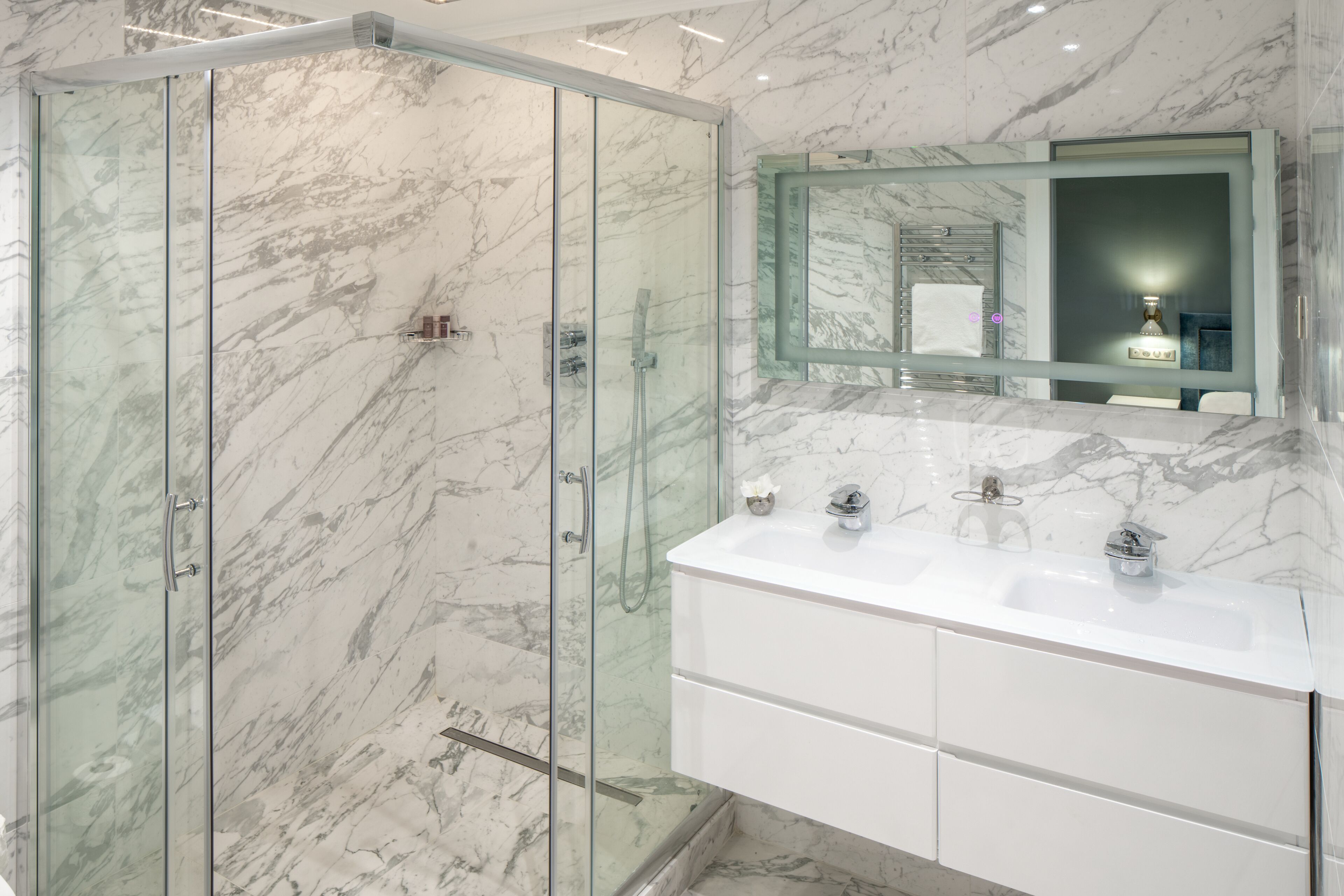 Une salle de bain moderne avec une douche à l’italienne en verre, un meuble-lavabo blanc doté d’un plan de toilette en marbre et un grand miroir au-dessus du lavabo.