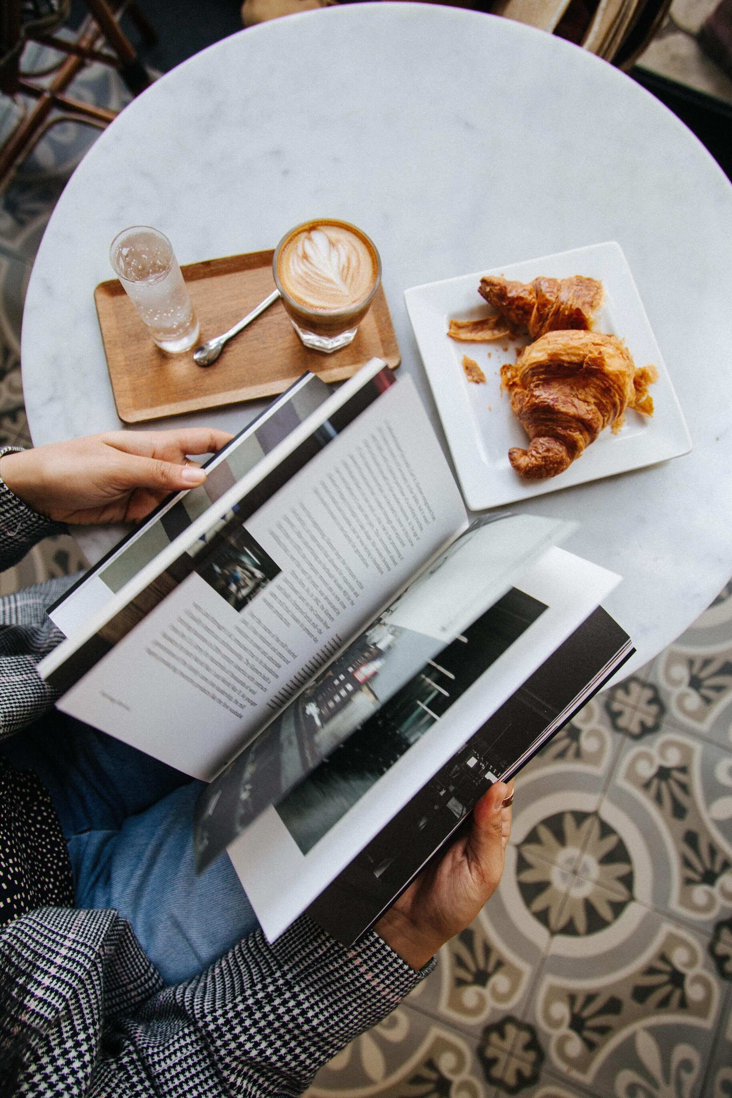 Una persona leyendo una revista en una mesa redonda, con un plato de croissants y una taza de café.