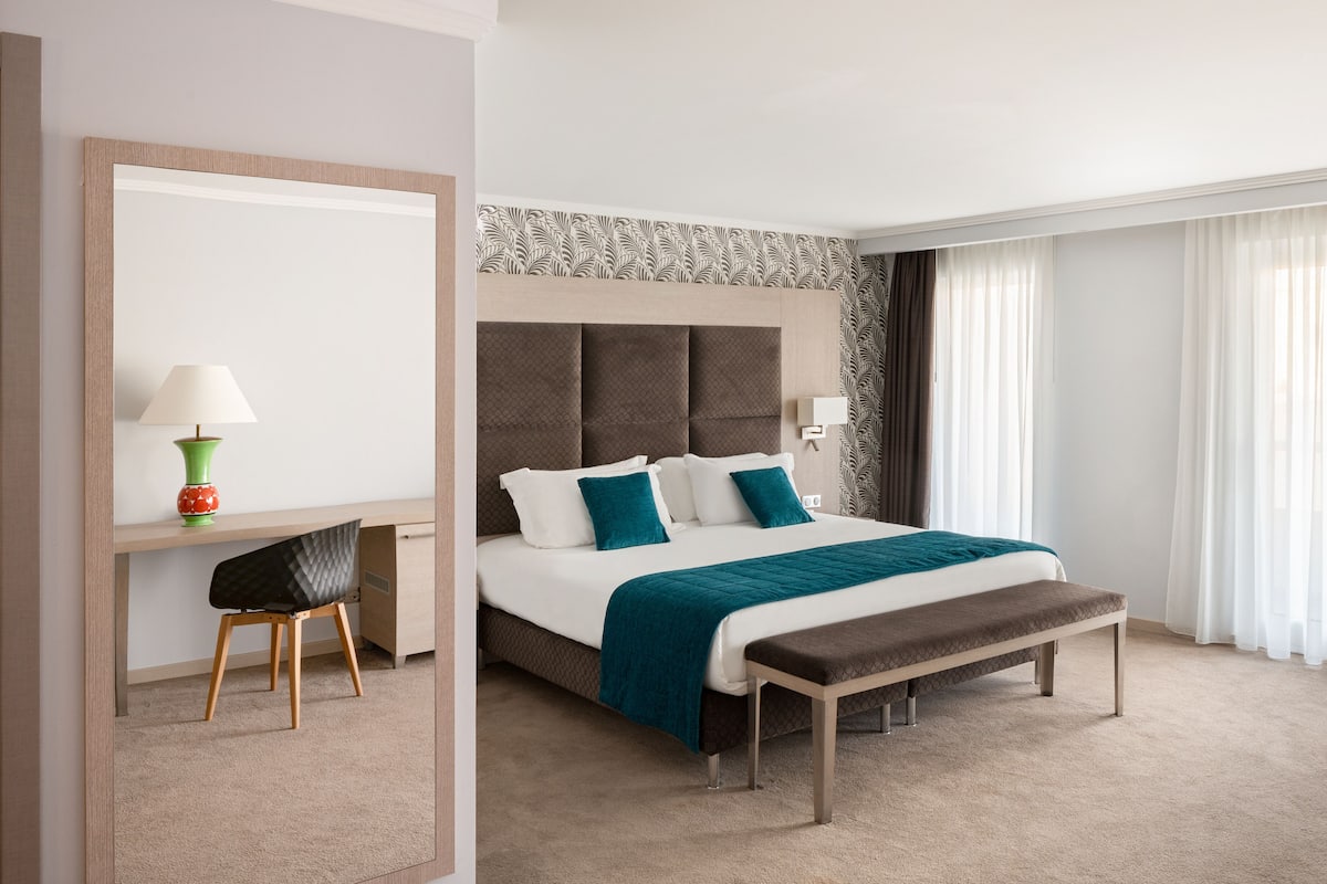 A hotel room with a bed, a desk, a chair, a bench, a lamp, and a large mirror.