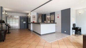 A modern reception area with a counter, a desk, and a chair. There are signs for elevators and stairs, and a wall-mounted light fixture. - Le Floréal (Vence)