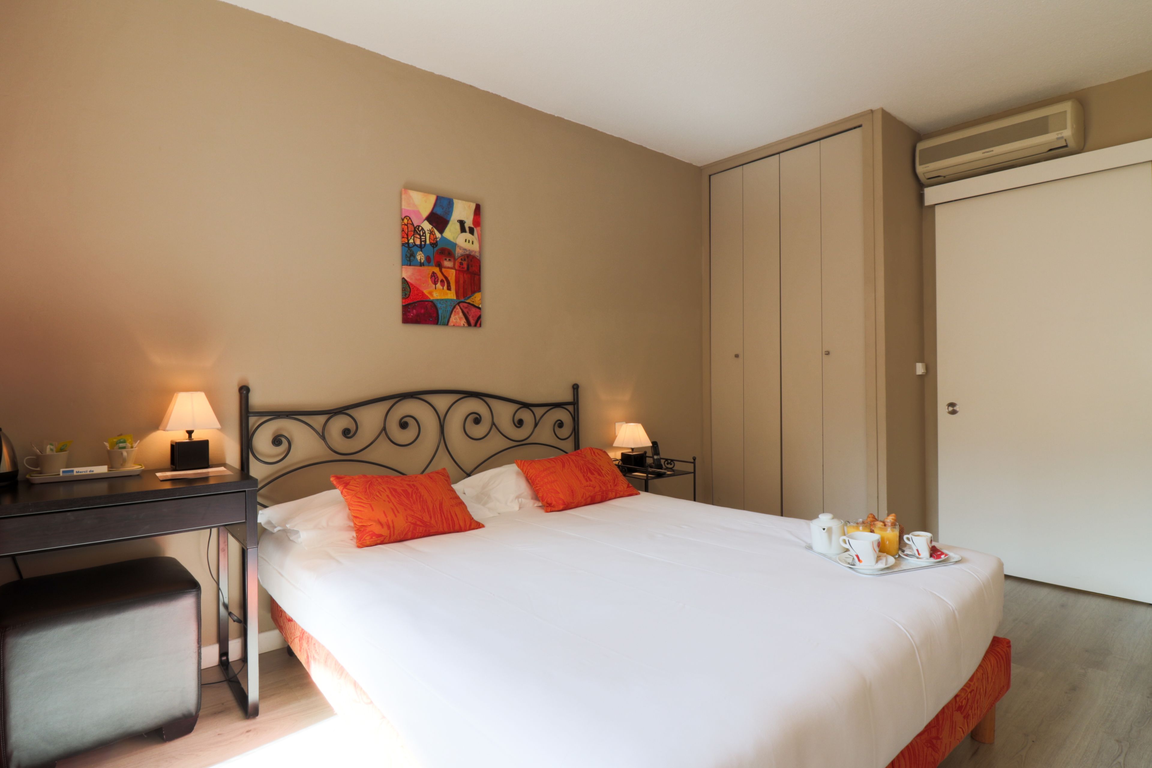 A neatly made bed with white and orange bedding, a desk with a lamp, and a small table with breakfast items.