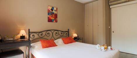 A neatly made bed with white and orange bedding, a desk with a lamp, and a small table with breakfast items.