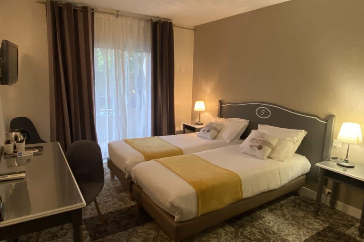 Une chambre d’hôtel avec deux lits, un bureau, une chaise, une lampe et une fenêtre avec des rideaux.