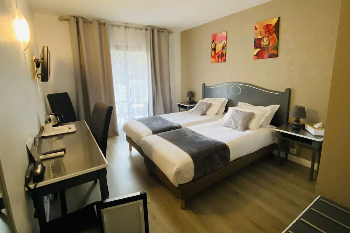 Une chambre d’hôtel avec deux lits, un bureau, une chaise, une lampe et des œuvres d’art accrochées aux murs.
