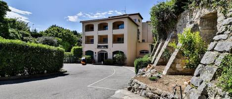 Un edificio a più piani con un ingresso centrale, circondato da vegetazione e un muro di sostegno in pietra.