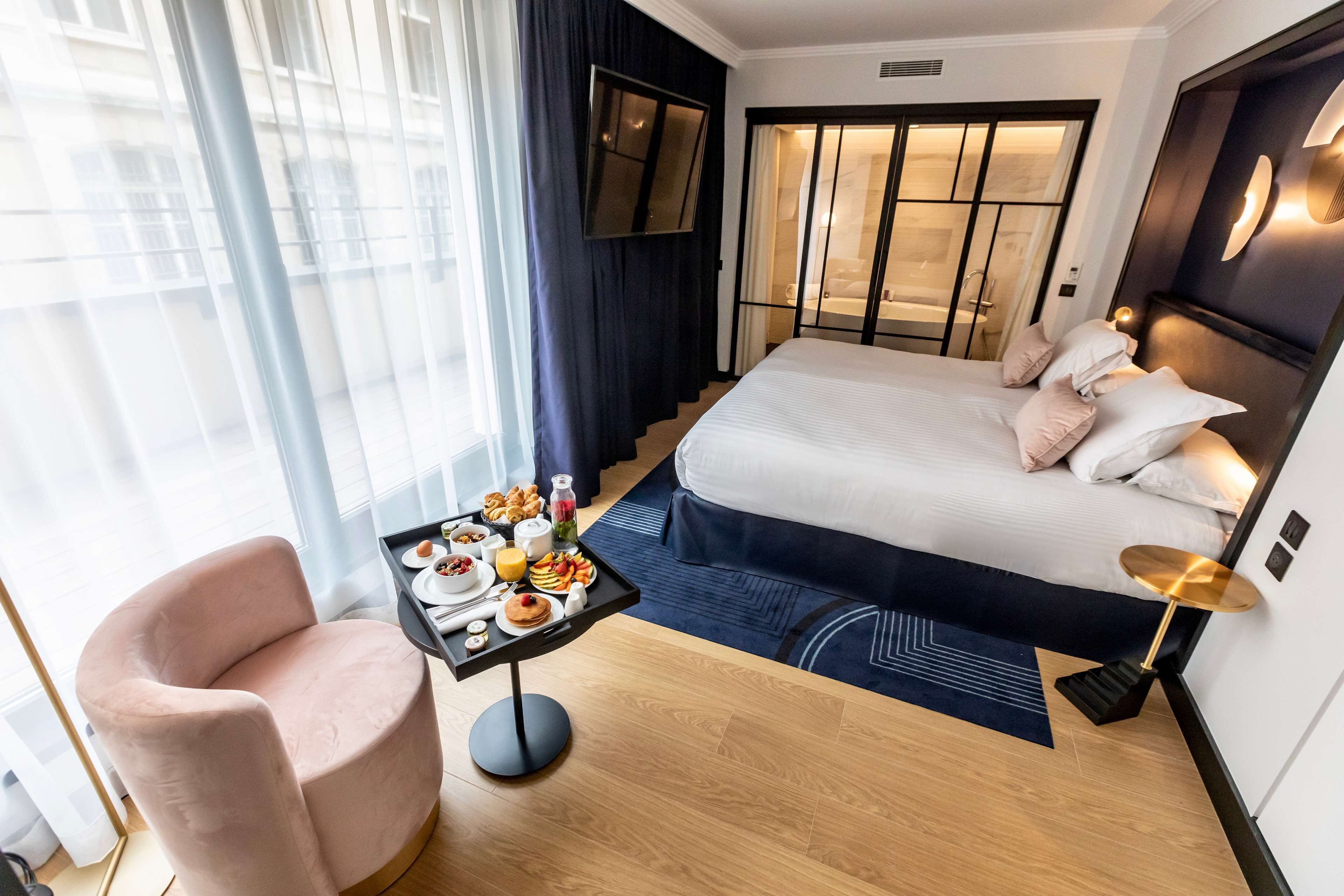 Habitación de hotel moderna con una cama grande, un escritorio con desayuno, una silla y vistas a la ciudad.