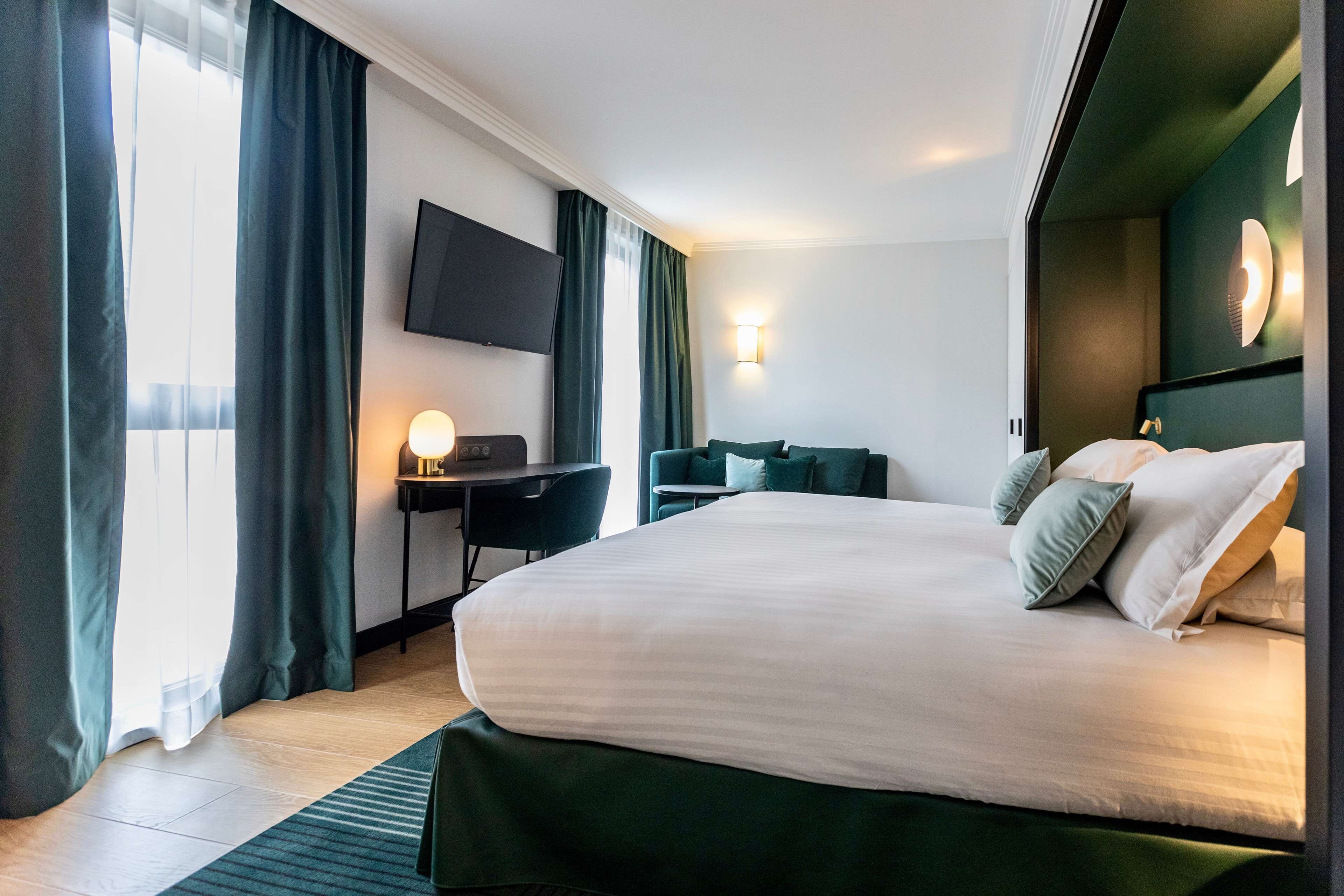 Une chambre d’hôtel avec un grand lit, un bureau avec une chaise, un téléviseur à écran plat fixé au mur et une fenêtre avec des rideaux.
