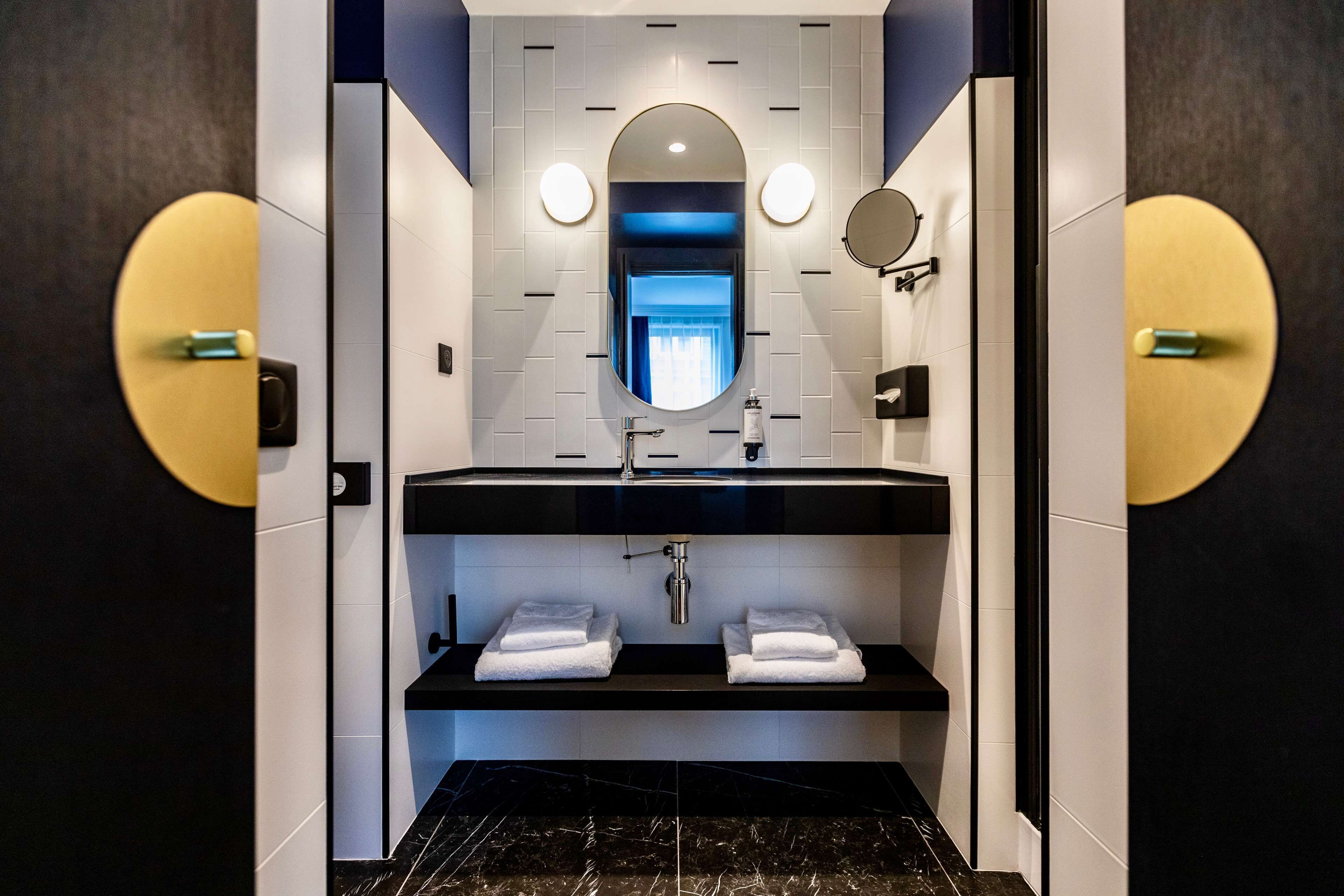 A modern bathroom with a black sink, white tiles, and a mirror above. There are two towel racks and a shelf below the sink.