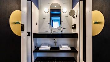 A modern bathroom with a black sink, white tiles, and a mirror above. There are two towel racks and a shelf below the sink.