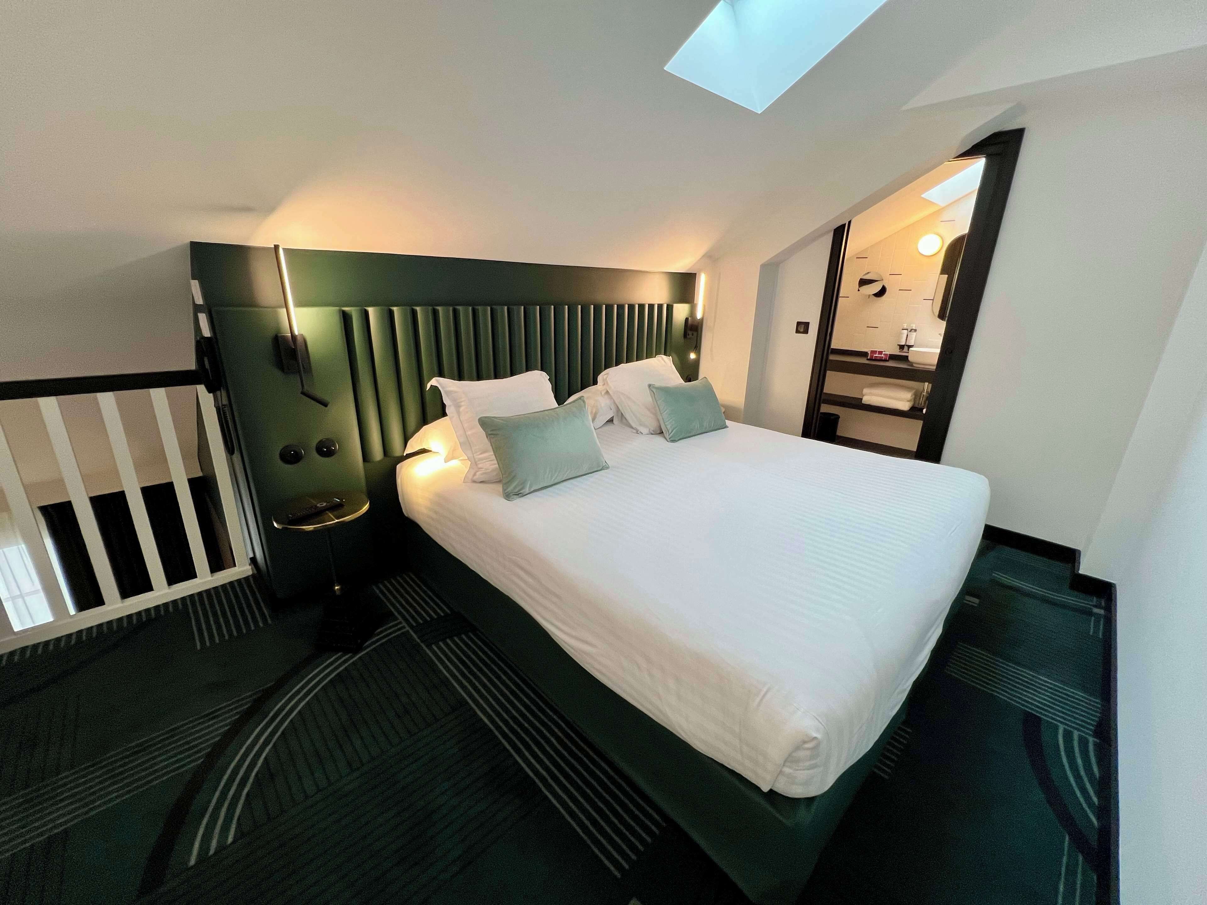 A modern hotel room with a large bed, a bedside table, a nightstand, a small window, and a mirror.