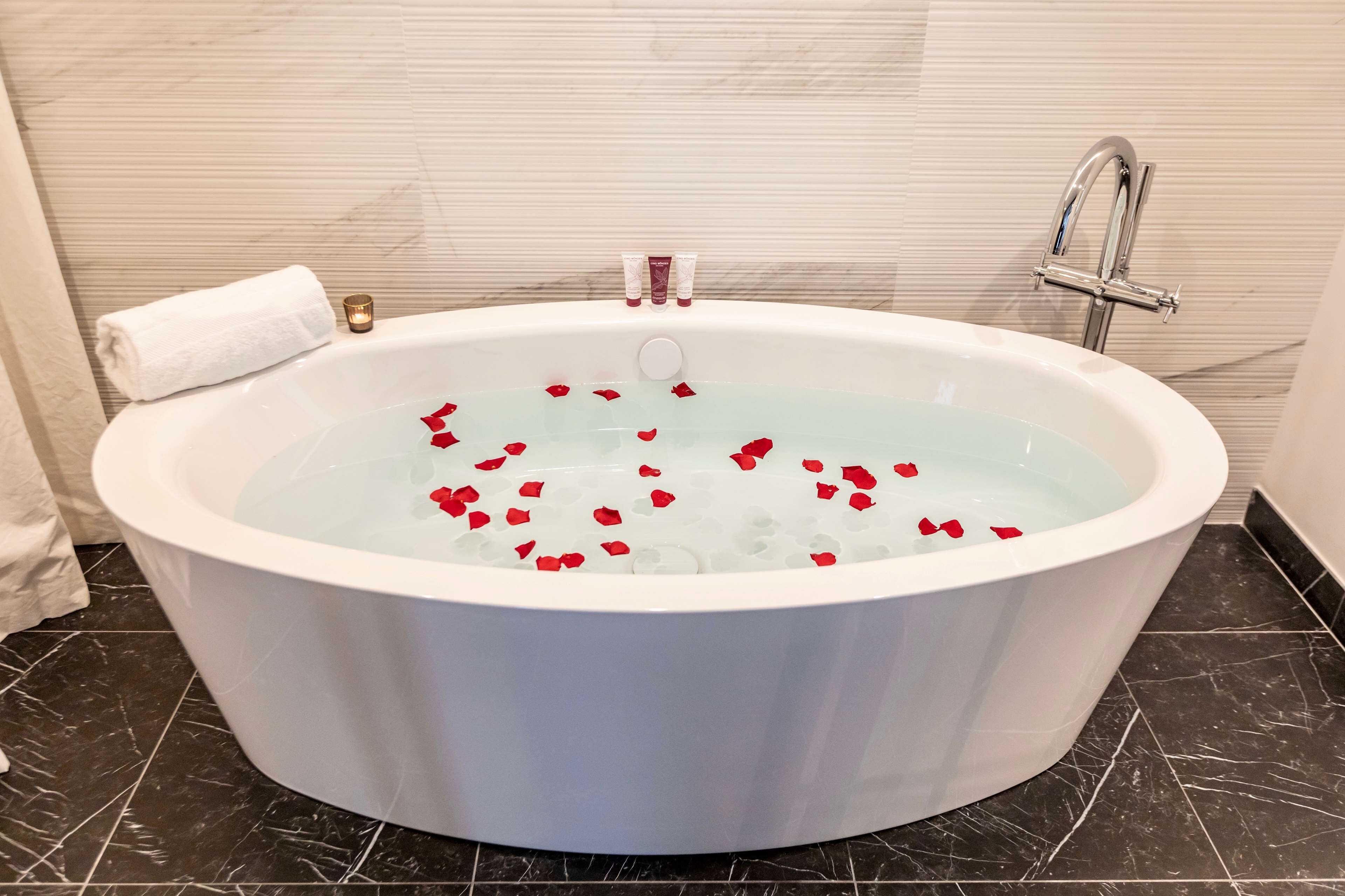 A white bathtub with rose petals floating in the water, a white towel, and a small candle on the side.