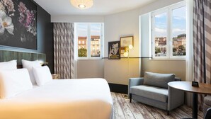 A hotel room with a large bed, a grey armchair, a small table, and a view of buildings through the windows.